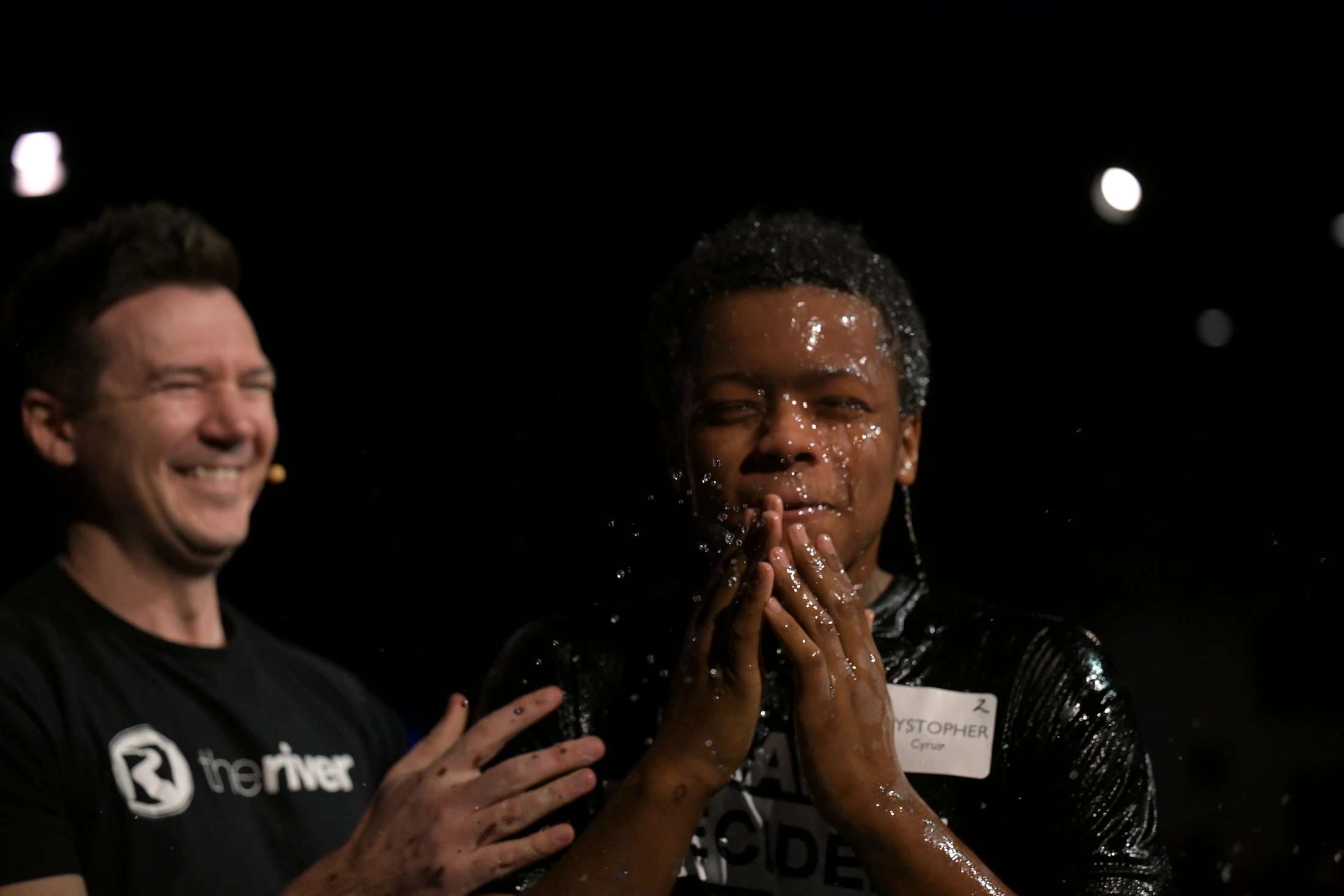 A young Black man with wet hair and face holds his hands in prayer after being baptized, while a smiling man in a 'the river' shirt looks on.